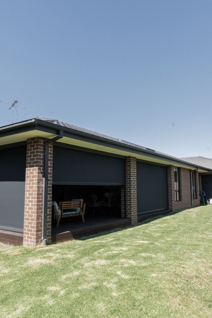 Run of motorized zip-track screens lowered across a brick ranch home patio with one bay open to seating